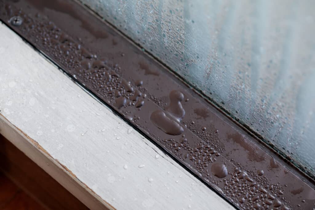 Gouttes d'eau de condensation sur un cadre de fenêtre marron et blanc, avec de l'humidité sur la vitre texturée.