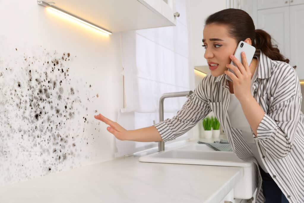 Femme désemparée téléphonant et pointant du doigt une large tache de moisissure noire sur le mur blanc d'une cuisine moderne.